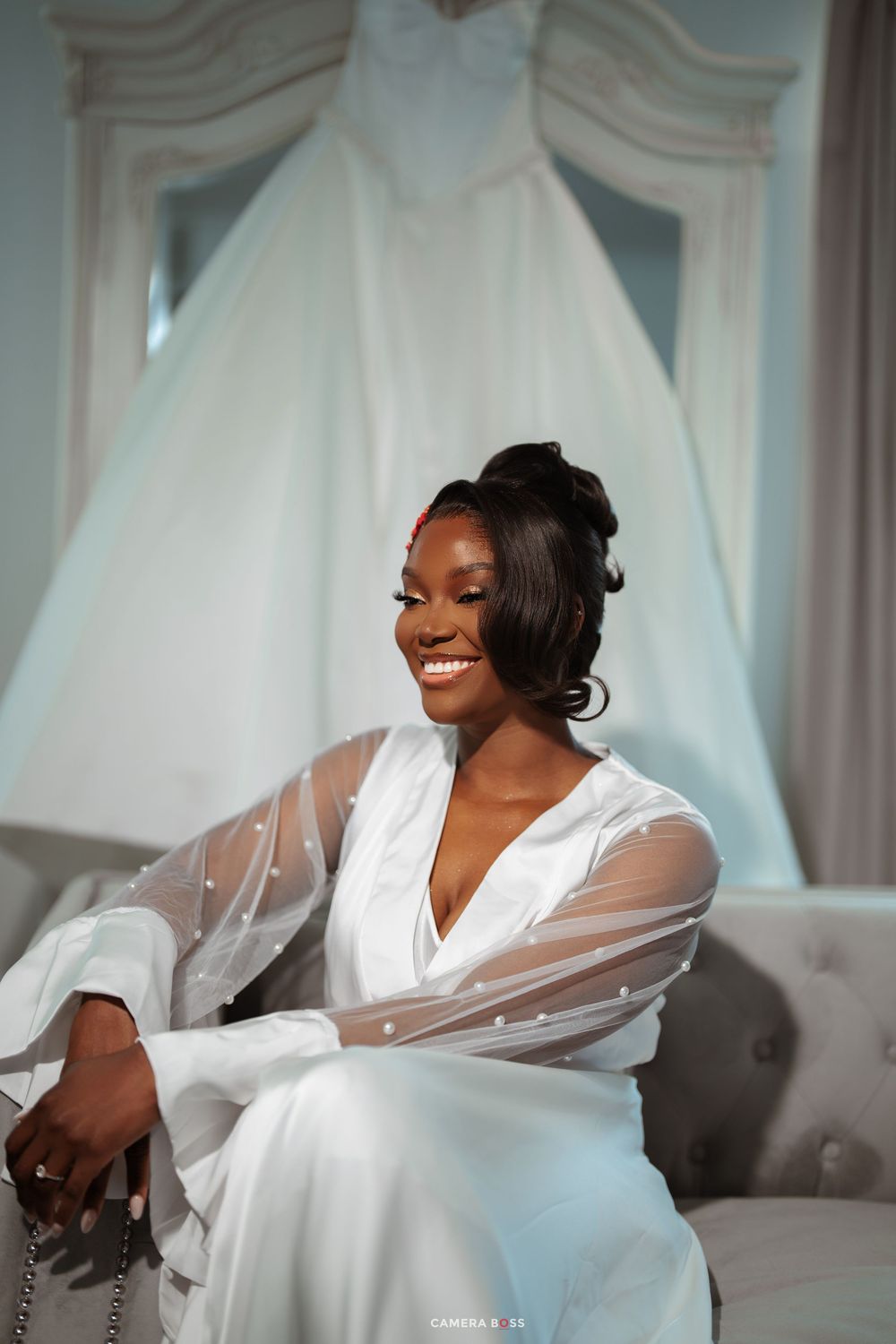 Smiling bride in white sheer robe sits elegantly with wedding gown displayed on hanger in bright bridal suite.