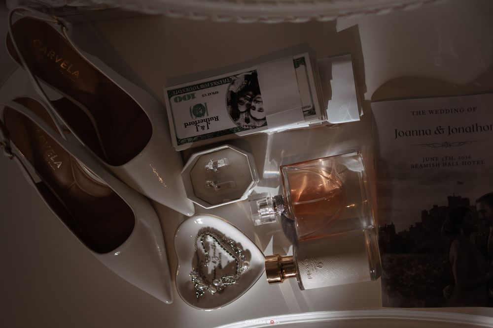 Flat lay of wedding accessories including heels, perfume bottle, jewelry, cash, and stationery on white surface.