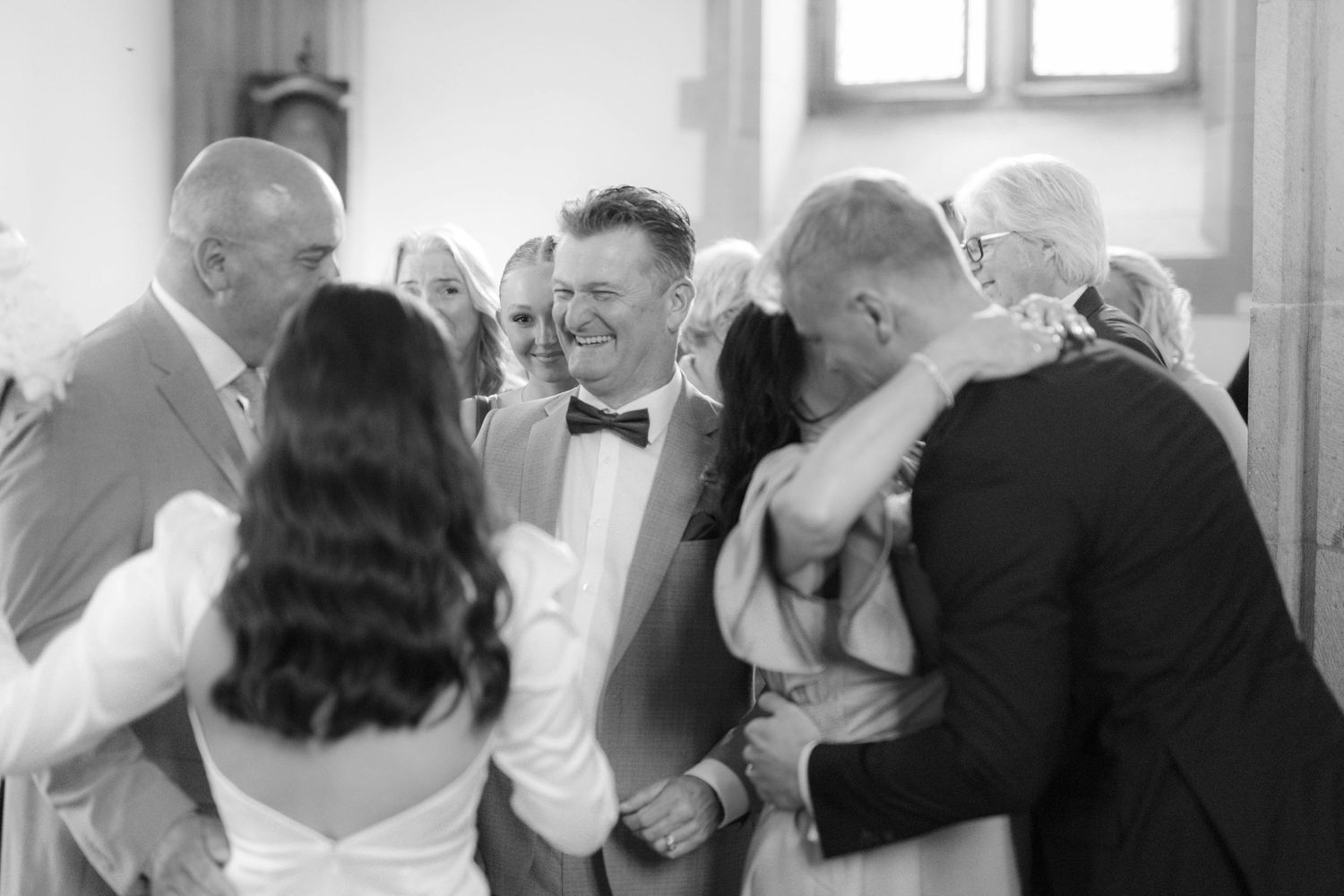 Black and white wedding photo of smiling couple holding hands surrounded by joyful guests in a church.