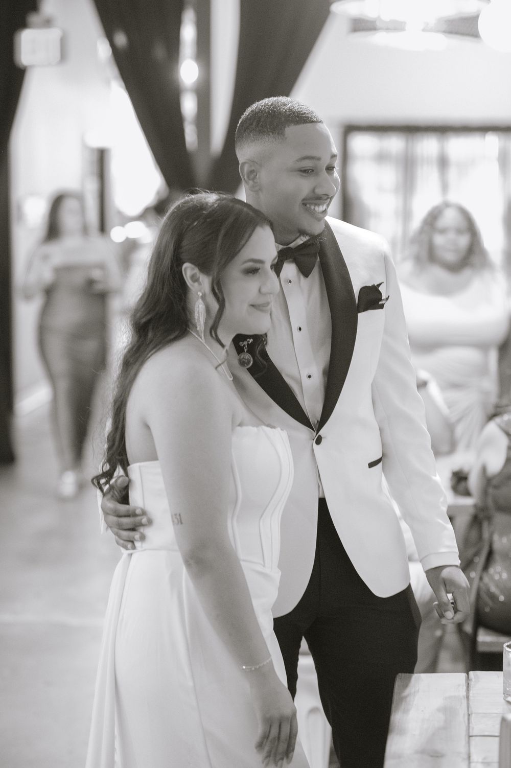Black and white photo of a smiling bride in white gown and groom in white tuxedo jacket at their wedding reception.