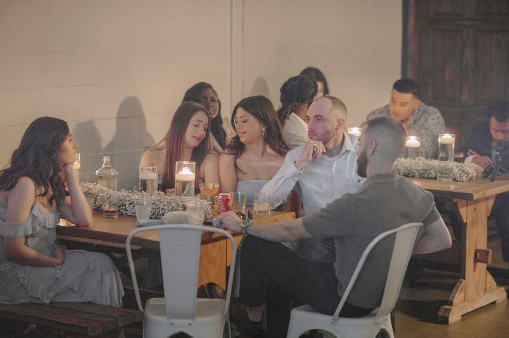 Group of elegantly dressed guests gathered around a candlelit wooden table at an intimate indoor event.