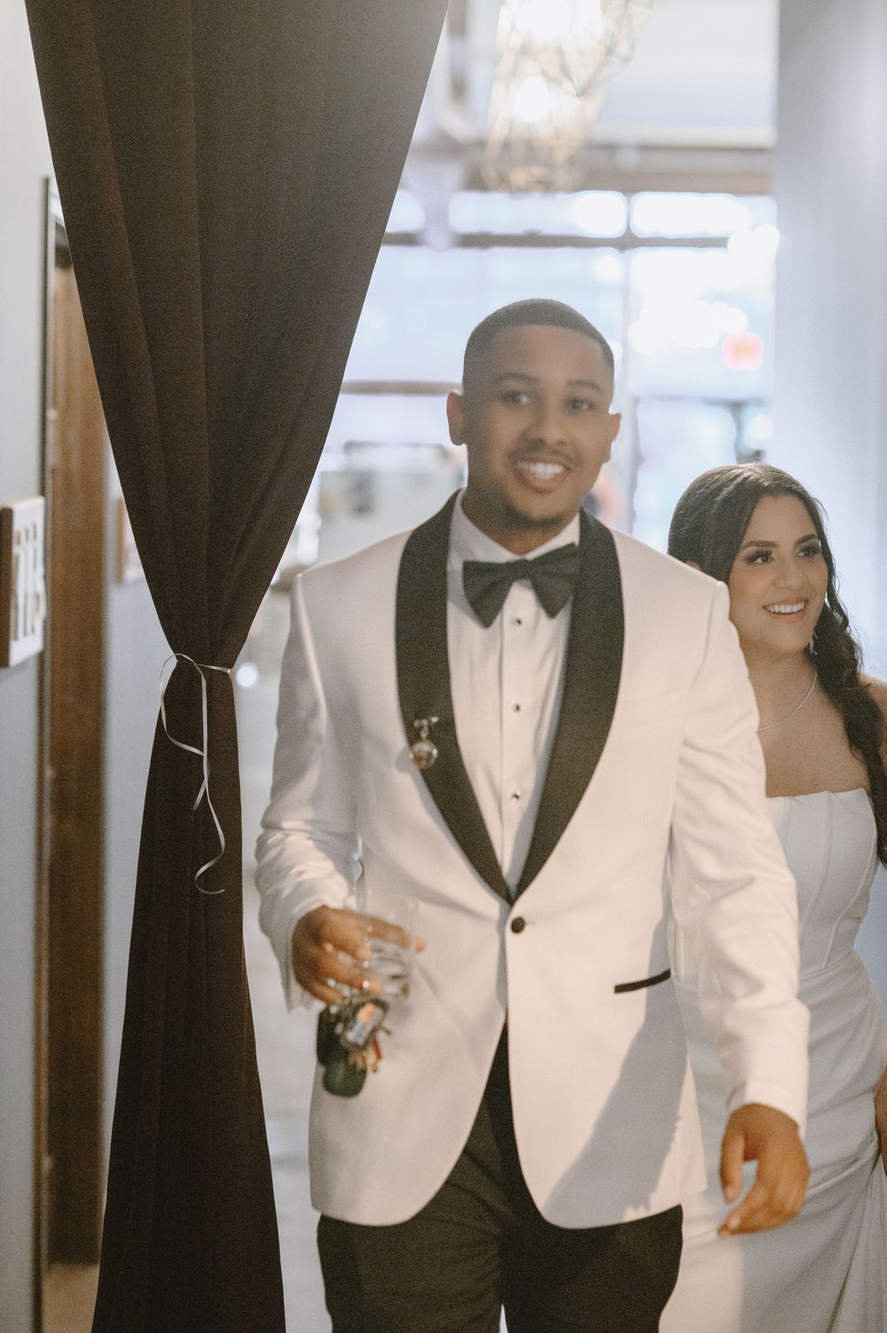 A smiling groom in a white tuxedo jacket with black lapels walks with his bride in a white gown at their wedding reception.