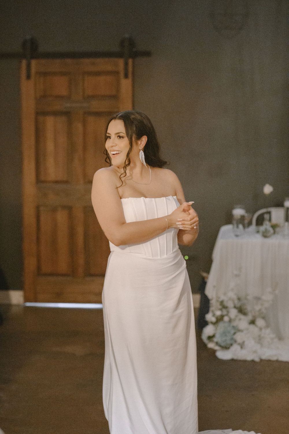 Bride in strapless white gown smiling during first dance at rustic wedding venue with wooden barn door backdrop.