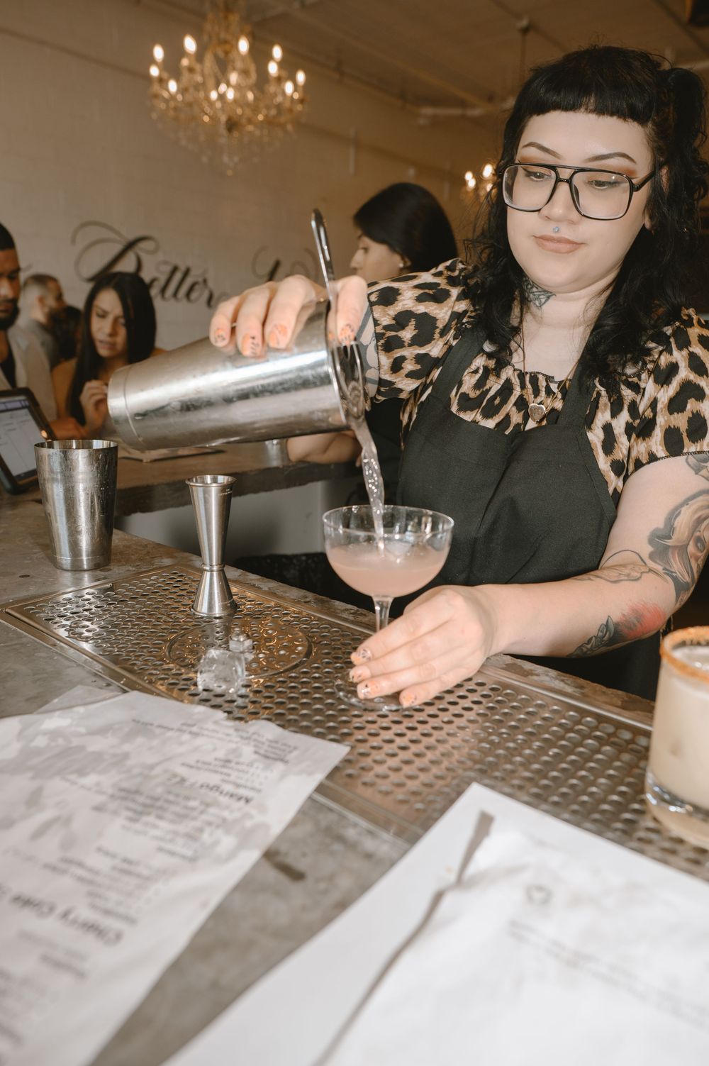 Tattooed female bartender with glasses pouring a pink cocktail into a coupe glass at an elegant bar.