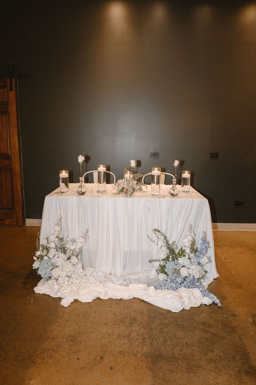 Elegant wedding sweetheart table with blue florals, candles, and flowing white linen draping.