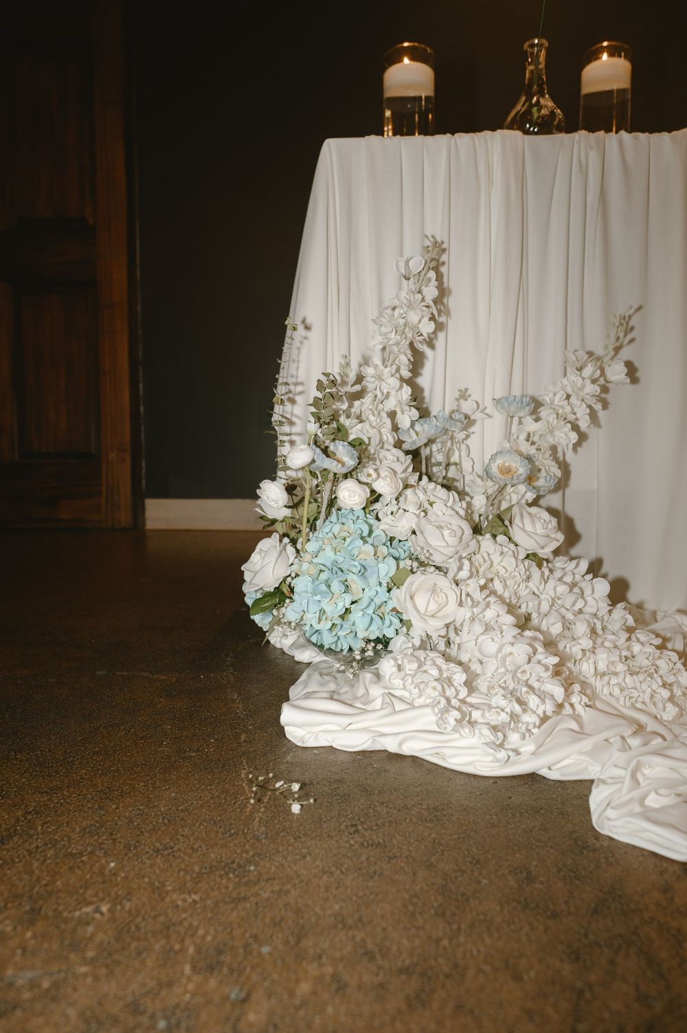 Elegant wedding table draped in white fabric adorned with white and blue hydrangea floral arrangement.