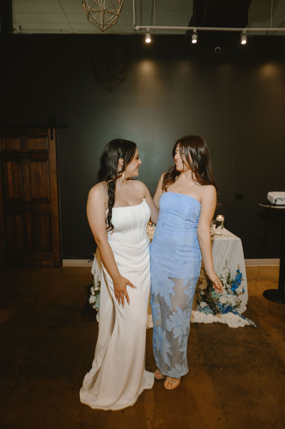 Bride in white strapless gown stands with bridesmaid in blue lace dress at elegant wedding reception venue.