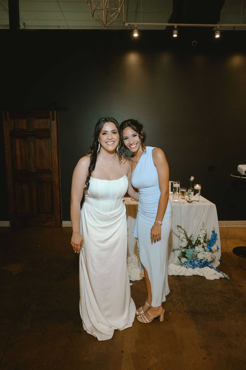 Bride in white gown poses with guest in blue jumpsuit at elegant wedding reception venue.