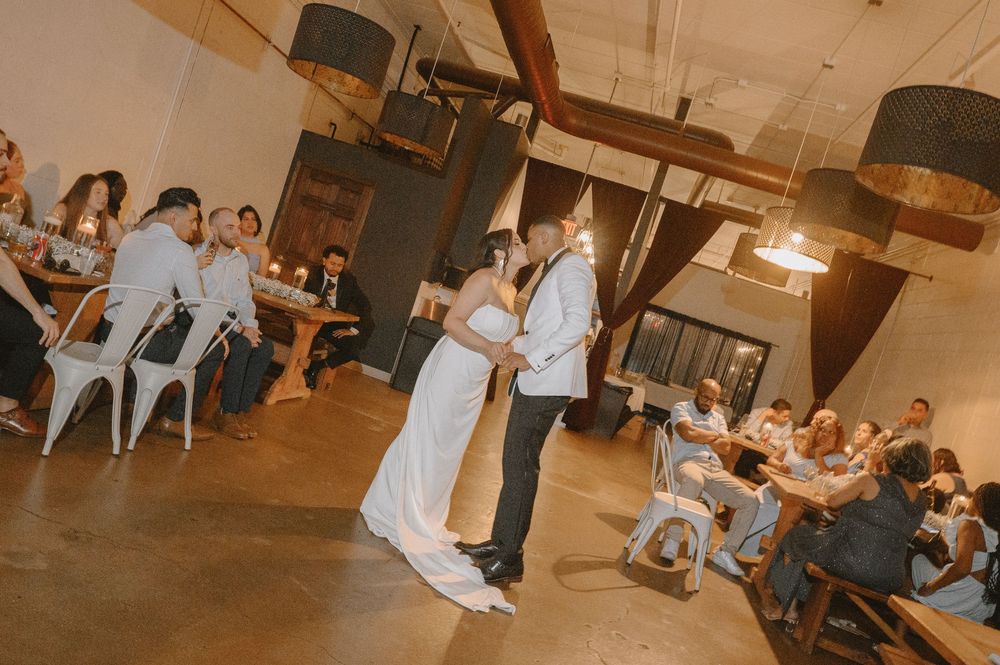Bride and groom share their first dance at a rustic wedding reception with guests seated around them.
