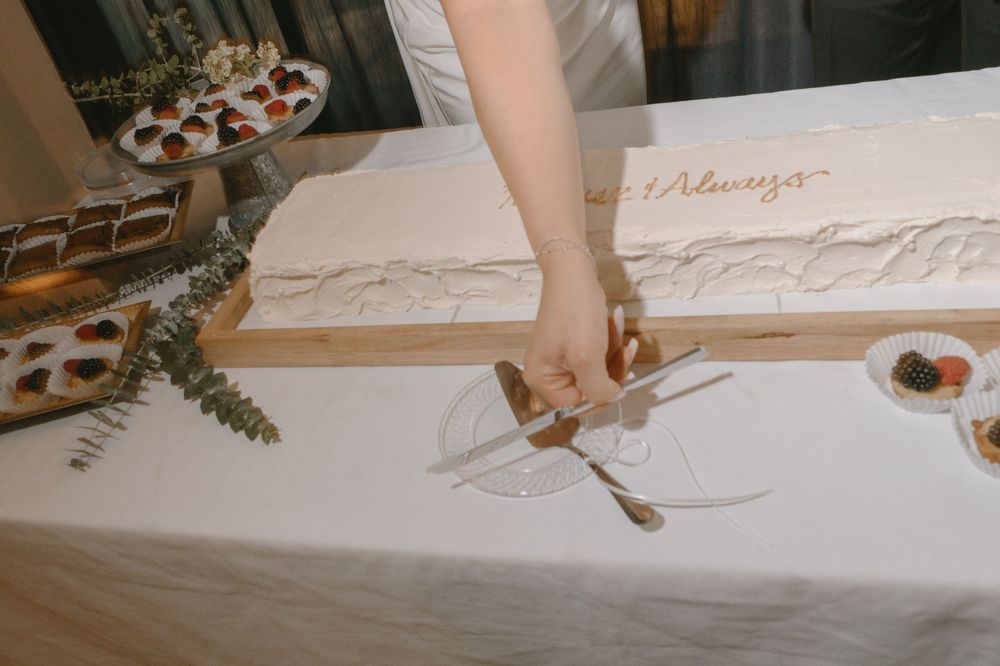 Bride cutting a elegant white wedding cake on a wooden board at a reception table with floral decorations.
