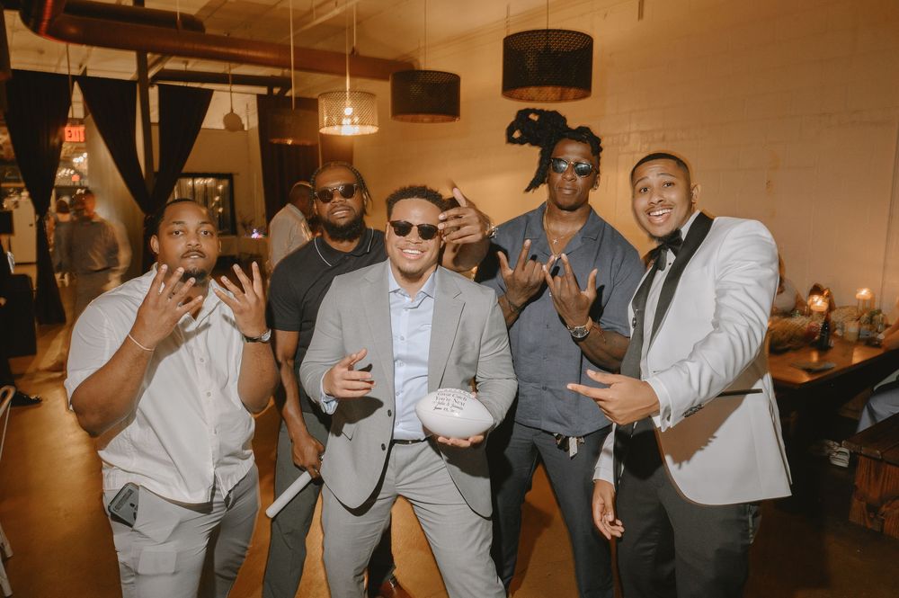 Five men in suits posing playfully at a wedding reception, one holding a white bouquet.