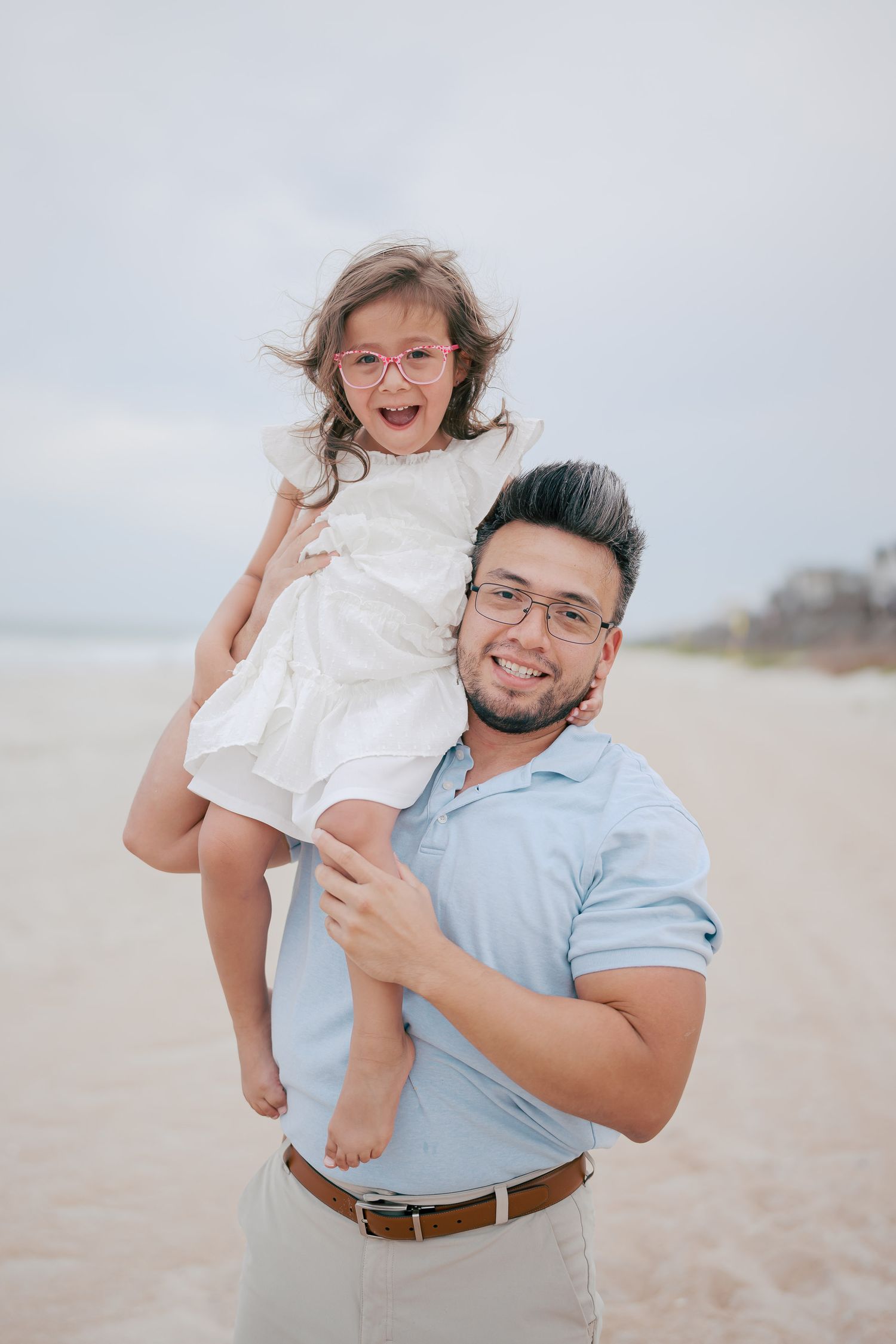 A smiling man in glasses holds a laughing little girl in a white dress on a beach.