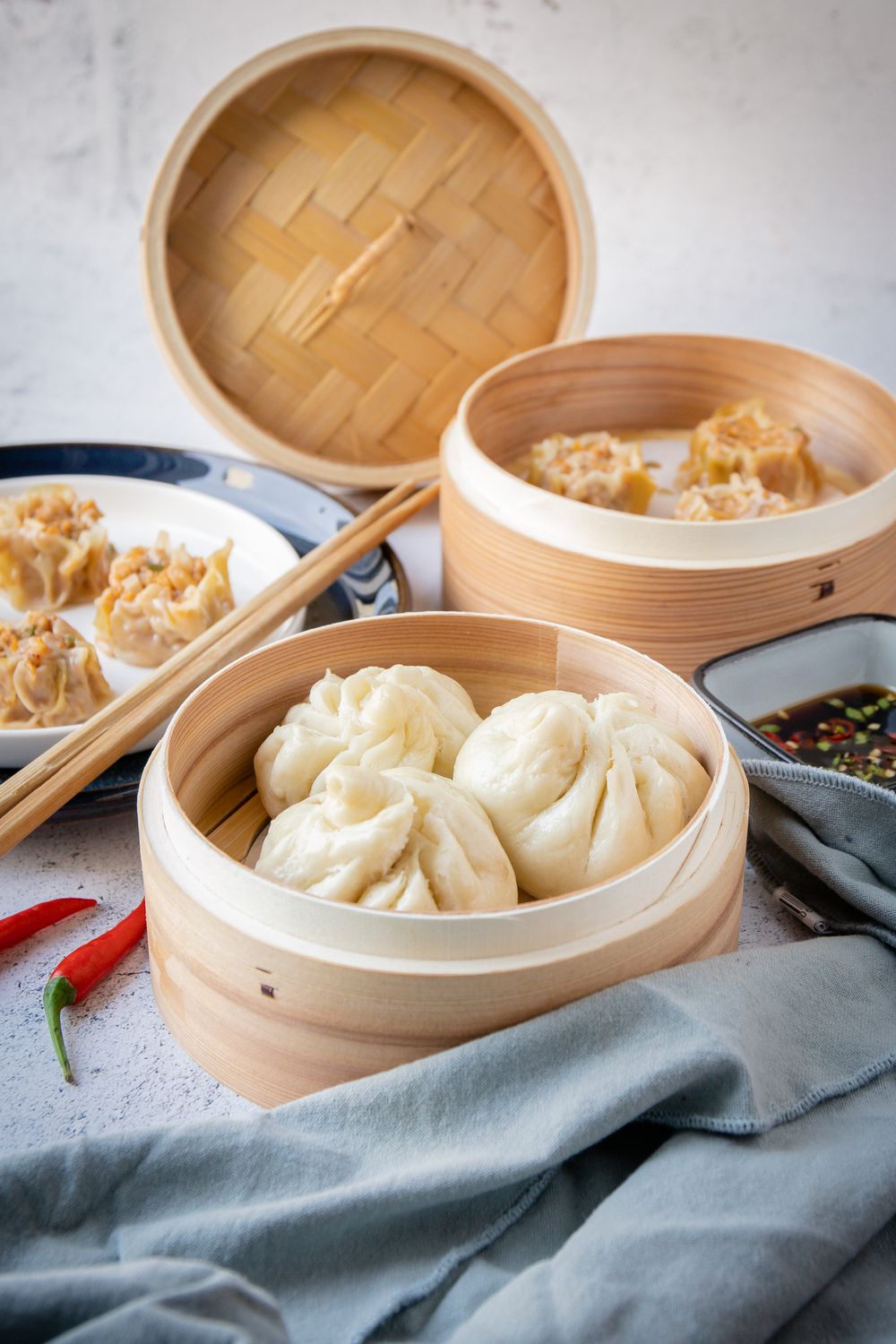 Steamed dumplings in bamboo baskets with dipping sauce and chopsticks on a gray linen cloth.