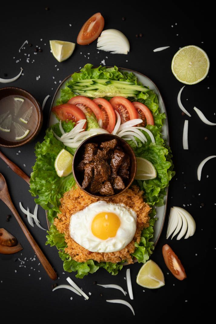 Nasi goreng topped with sunny side up egg, mushrooms, lettuce and lime wedges served on dark plate with onions.