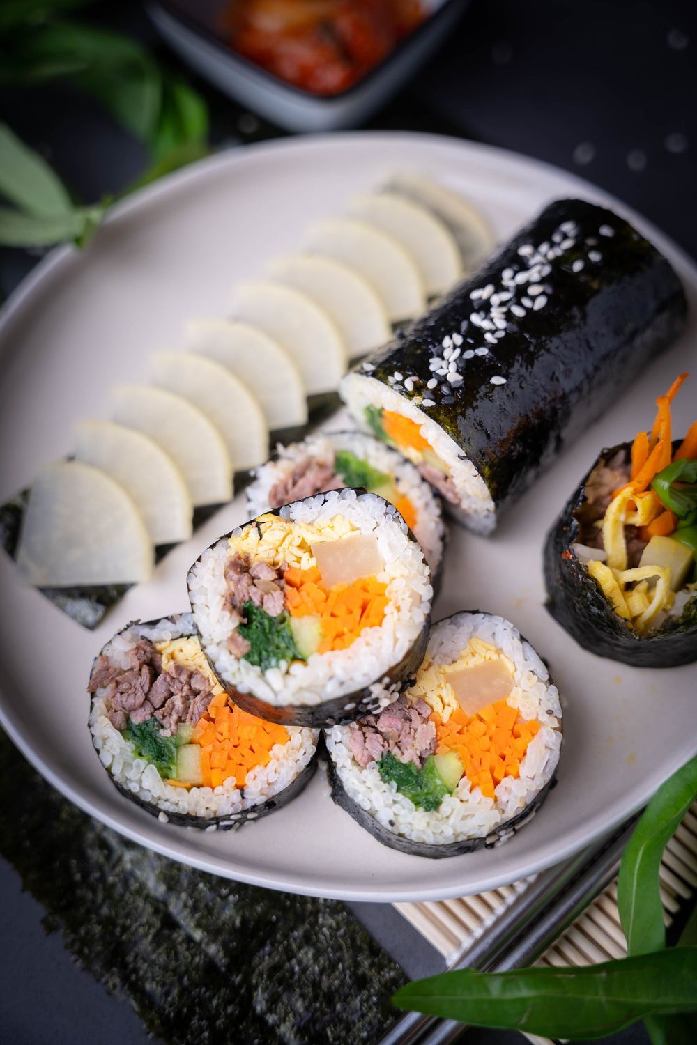Homemade gimbap rolls with rice, vegetables, and fish roe arranged on white plates with bamboo mats and garnish.