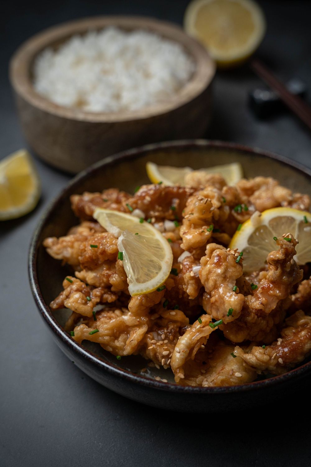 Crispy lemon chicken served on dark plates with rice and garnish in moody lighting.