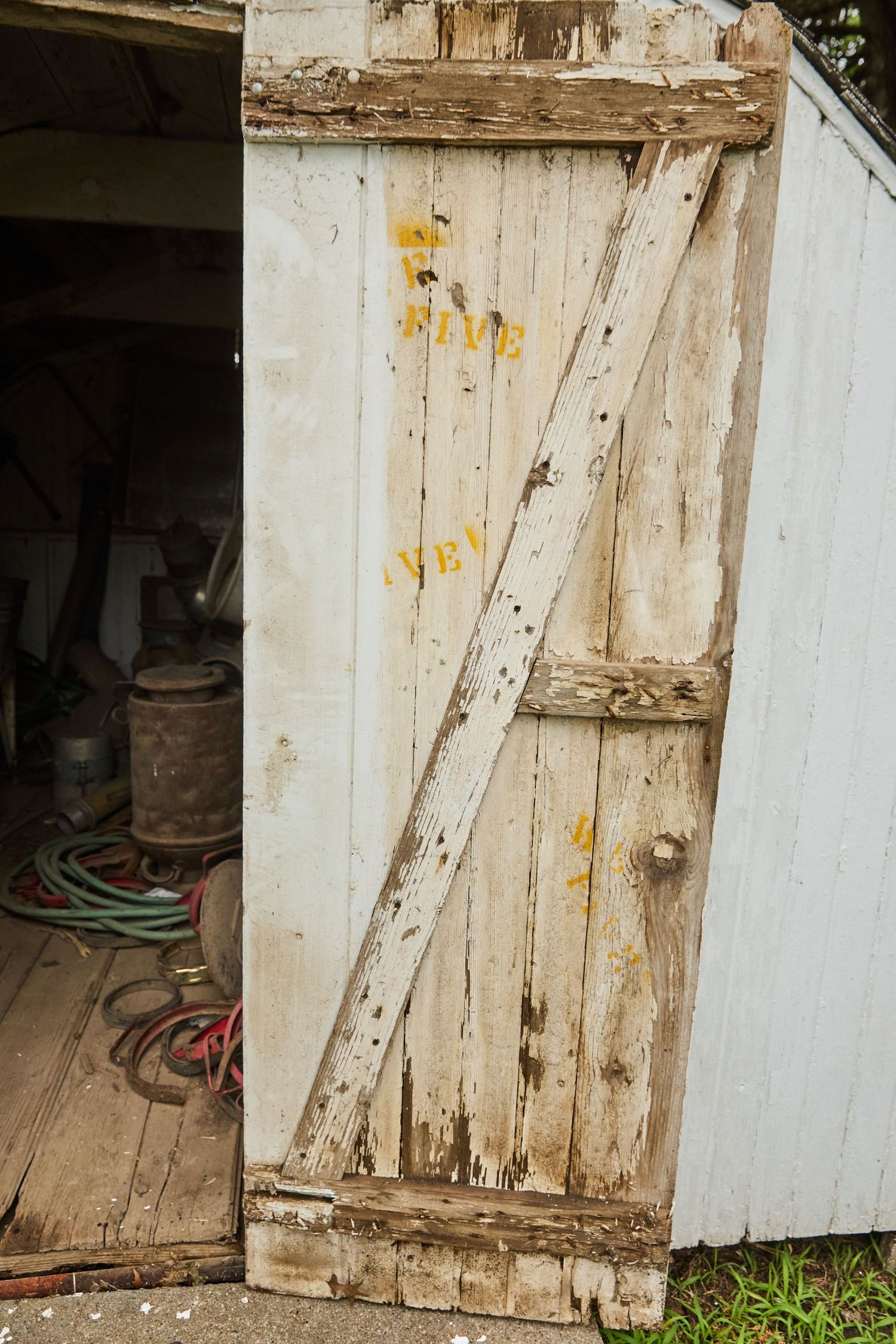Weathered white wooden shed door with peeling paint and yellow stenciled letters, slightly ajar revealing garden tools inside.