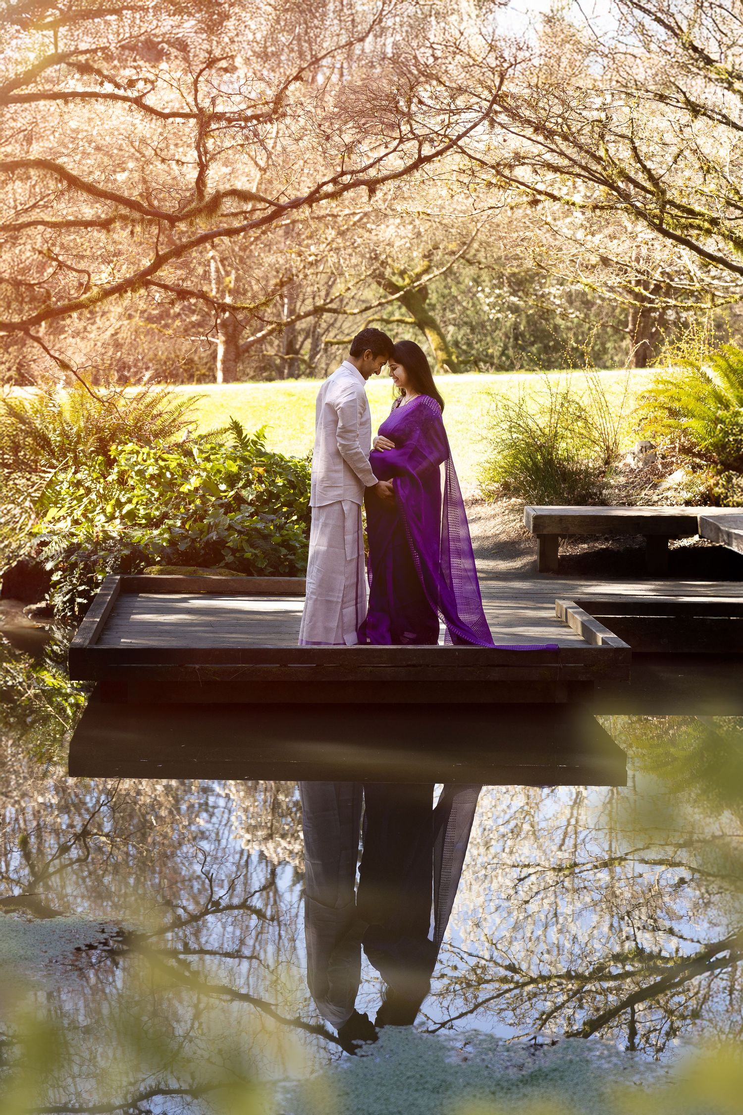 Woman in purple saree poses among white cherry blossoms in bloom, purple fabric flowing gracefully in a lush green park.