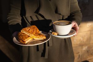 Person holding a plate with a croissant sandwich and a cup of latte coffee on a saucer in warm sunlight.