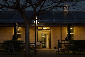 A stone ranch house with solar panels on the roof glows warmly at night, illuminated by outdoor lights.