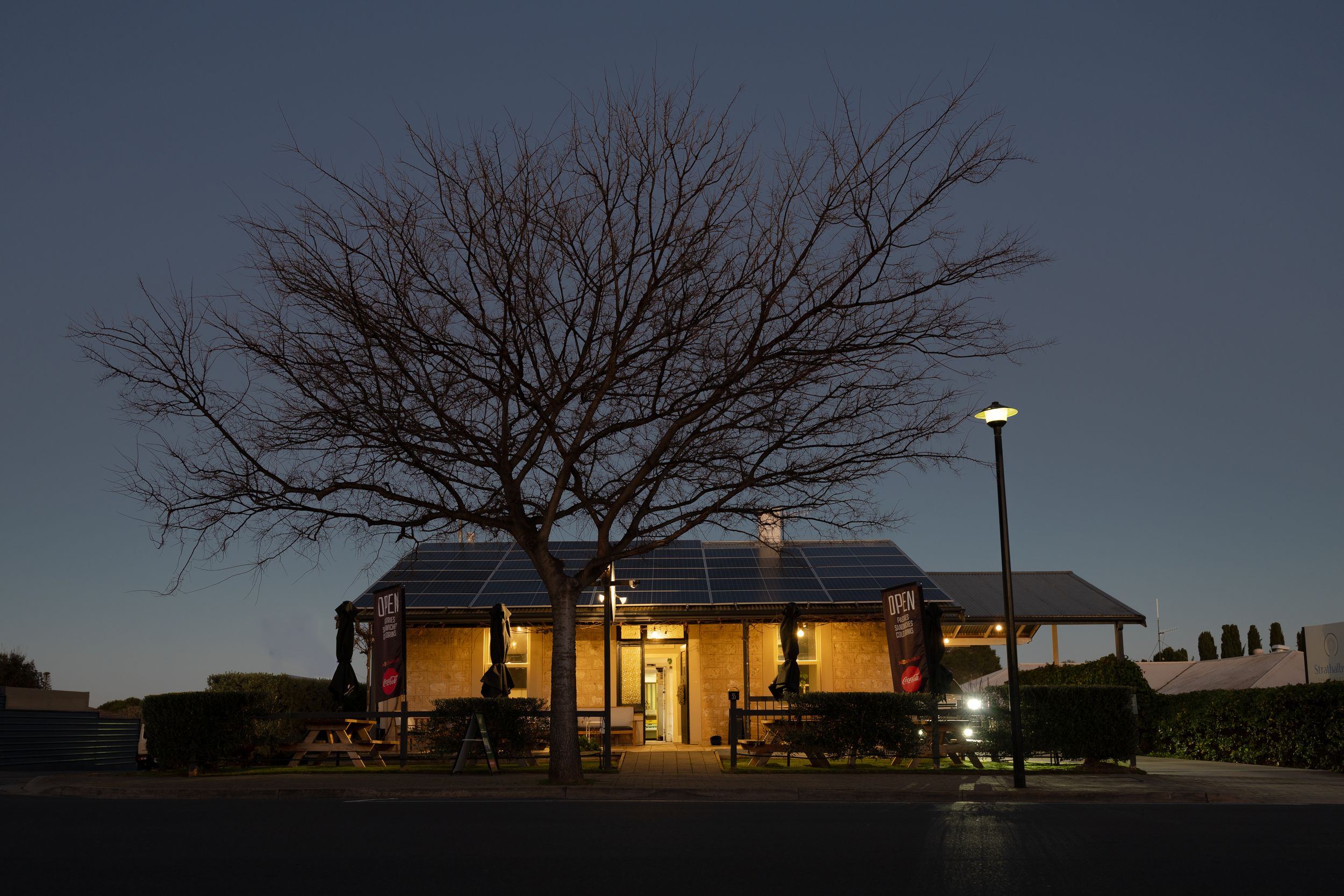 A cozy lit building glows warmly at dusk beneath a bare winter tree in a quiet parking lot.