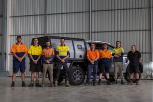 Team of seven workers in hi-vis uniforms posing in front of a ute inside a large industrial shed.