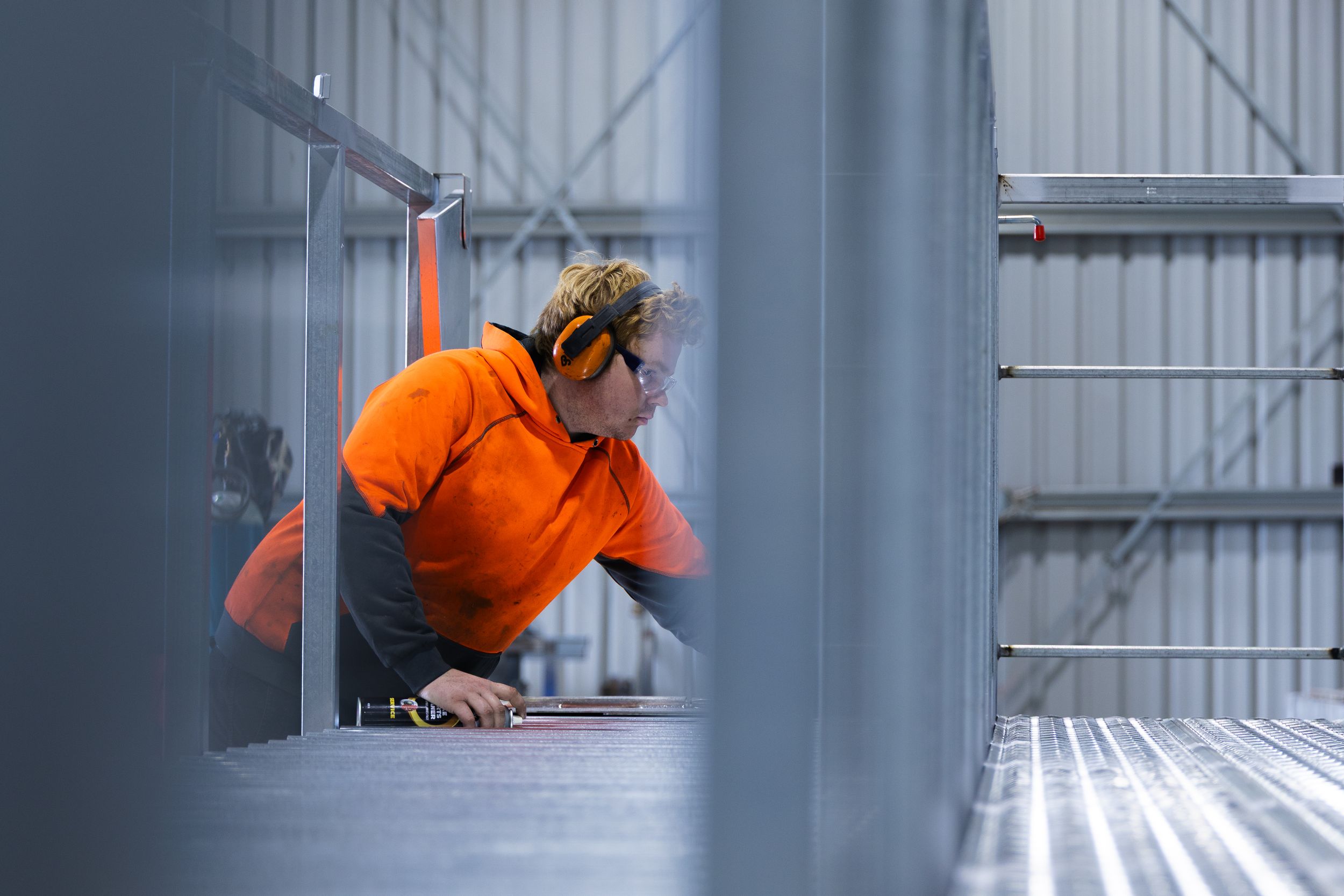 Worker in orange hi-vis shirt and ear protection inspects metal grating in a steel fabrication facility.