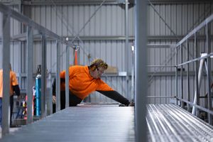 Worker in orange hi-vis gear carefully examines steel grating inside a metal fabrication warehouse.