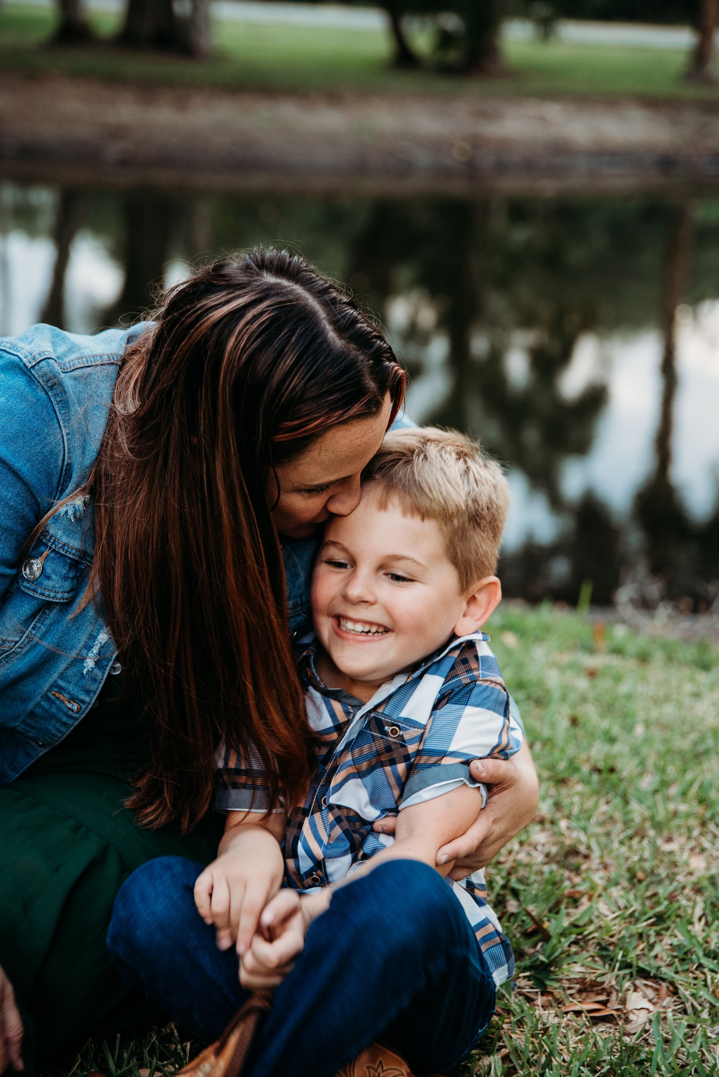 A tender moment shared between parent and child sitting by a pond on a grassy bank, both wearing blue.