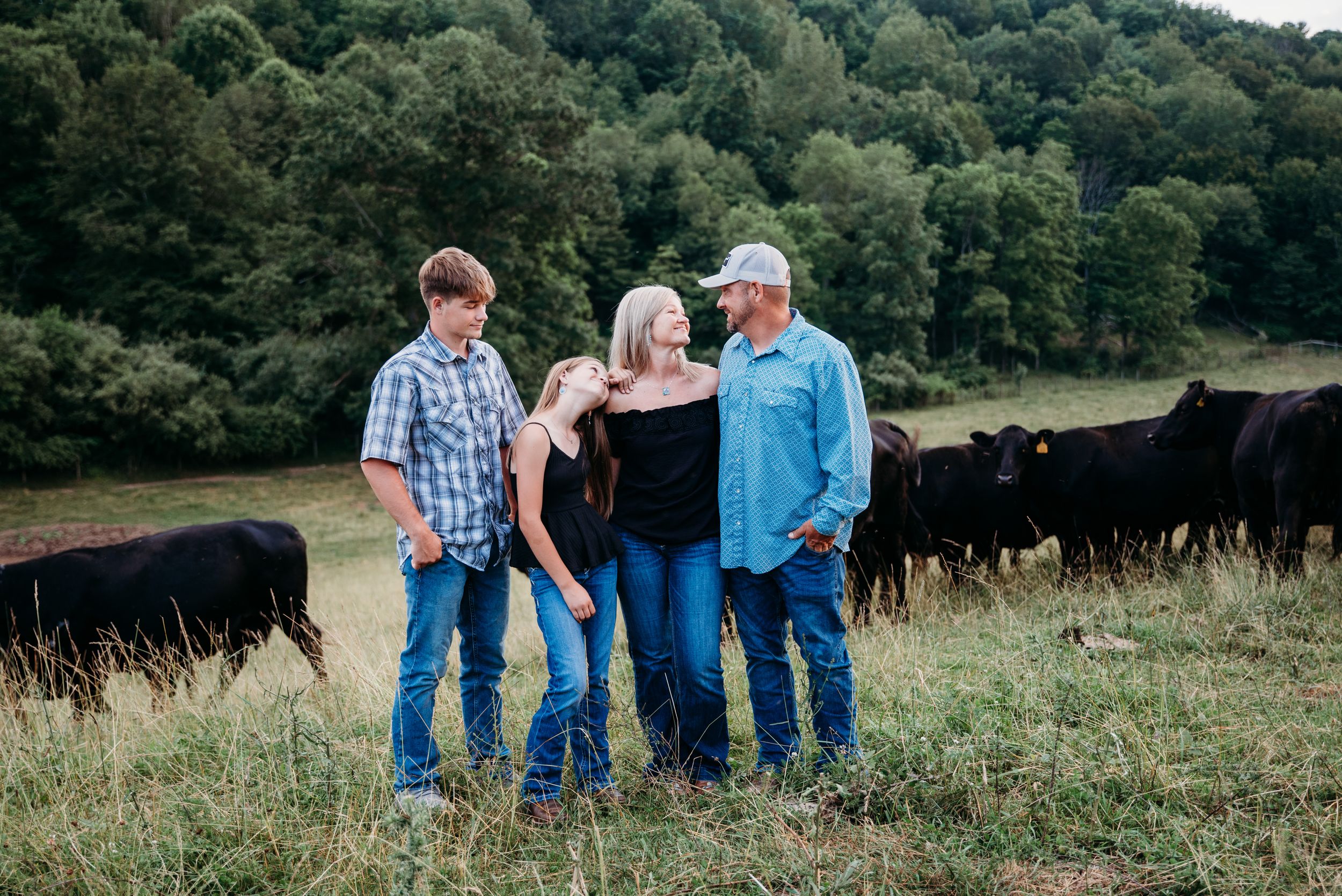 Three people stand together in a grassy field with black cattle grazing in the background near a wooded area.