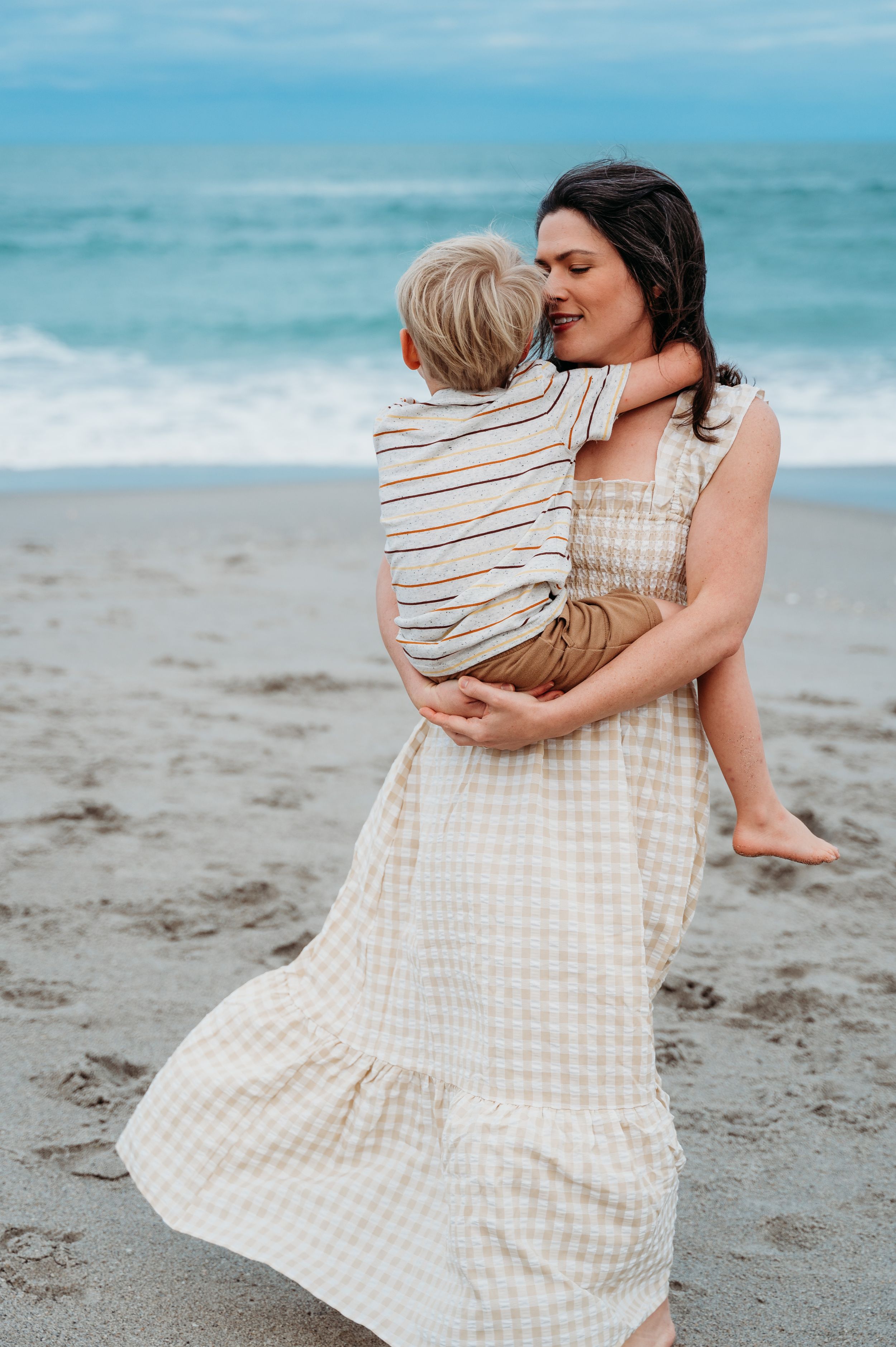 A tender moment captured at the beach with a beige gingham dress flowing in the ocean breeze.