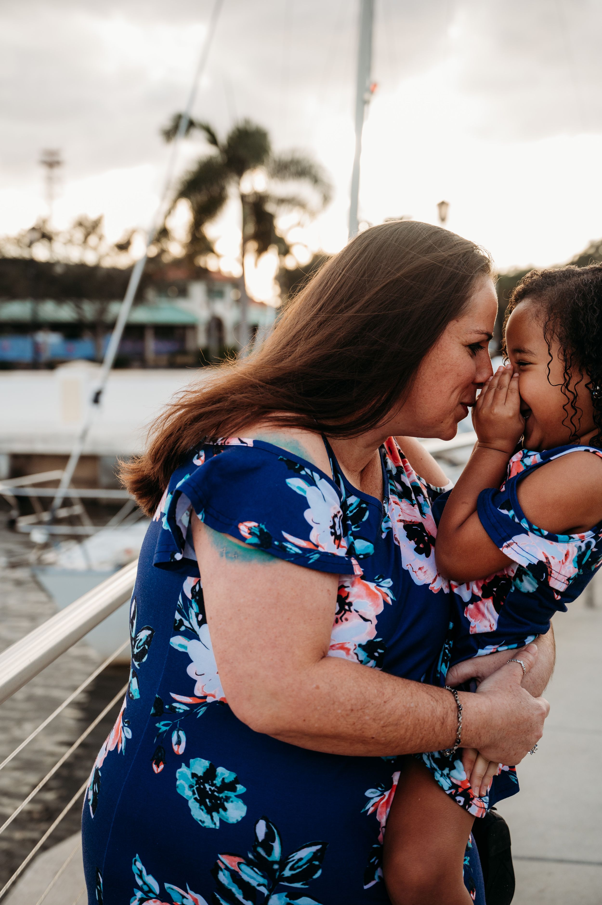 A tender moment shared between two people wearing matching blue floral dresses on a marina dock at sunset.