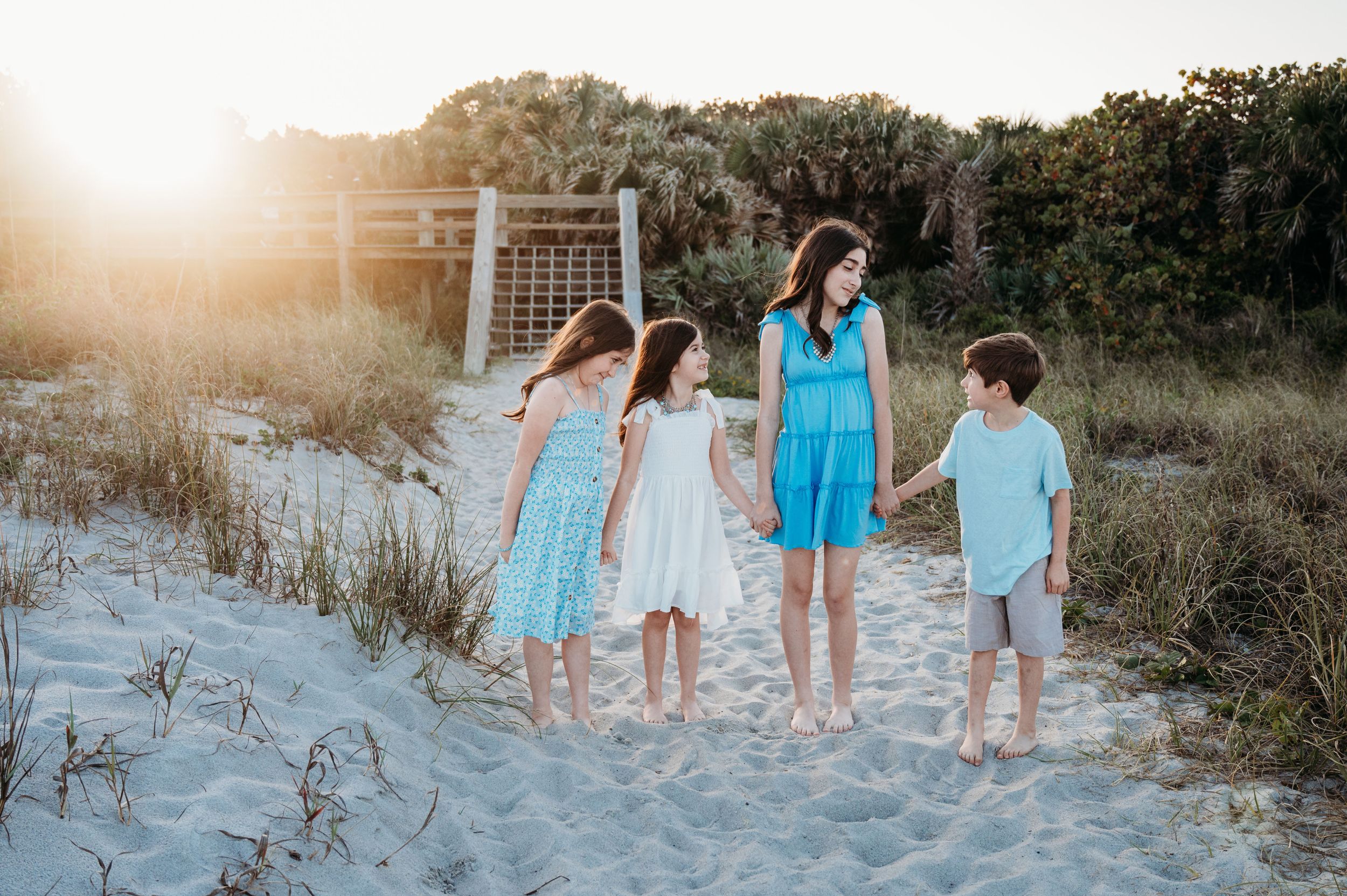 A group stands together on a sandy beach at sunset with tall grass and trees in the background.
