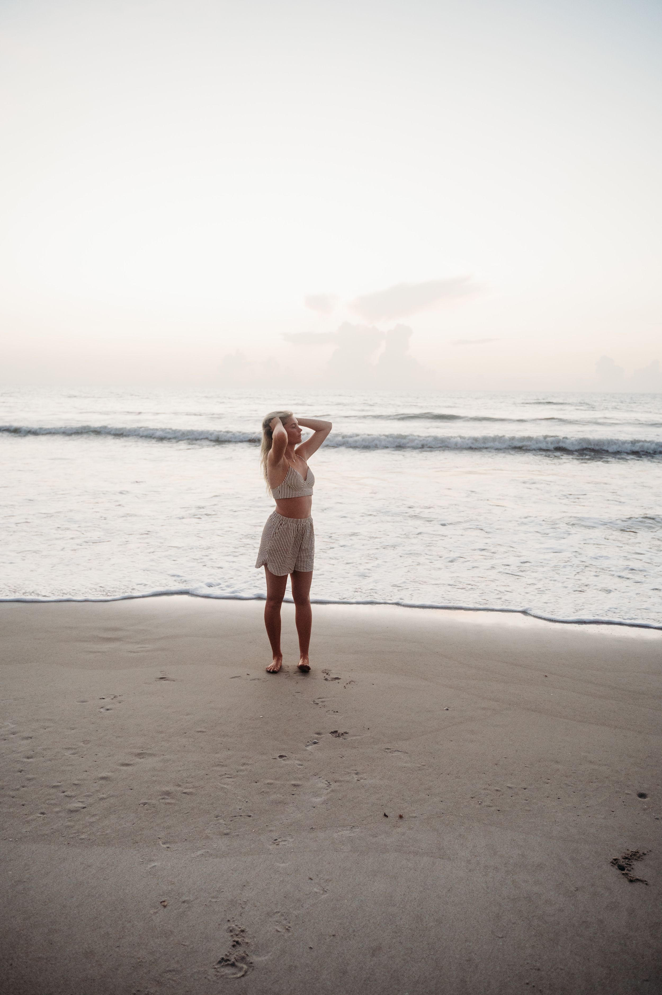 A person stands alone on a peaceful beach at sunset, watching the waves roll in while wearing casual summer attire.