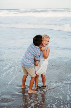 Two children share a playful moment while standing barefoot in the ocean waves at the beach.