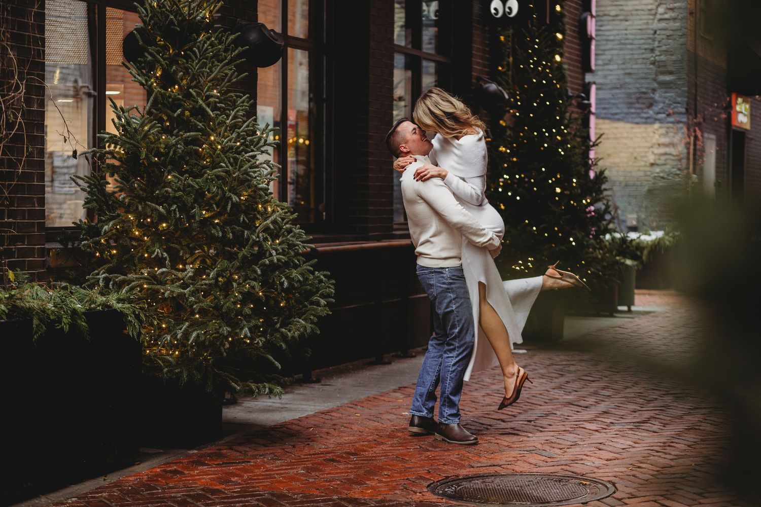 Couple embracing as a man lifts a woman in a white dress on a brick path lined with twinkling Christmas trees.