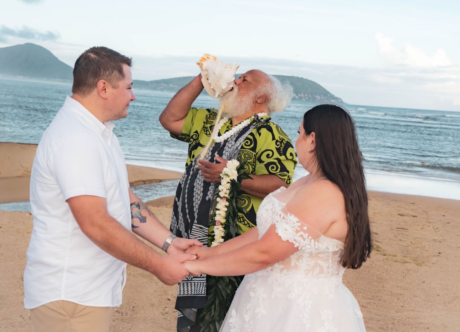 Bride in a white lace gown and groom in khaki shorts hold hands on a beach as an officiant in a green aloha shirt blows a conch shell.