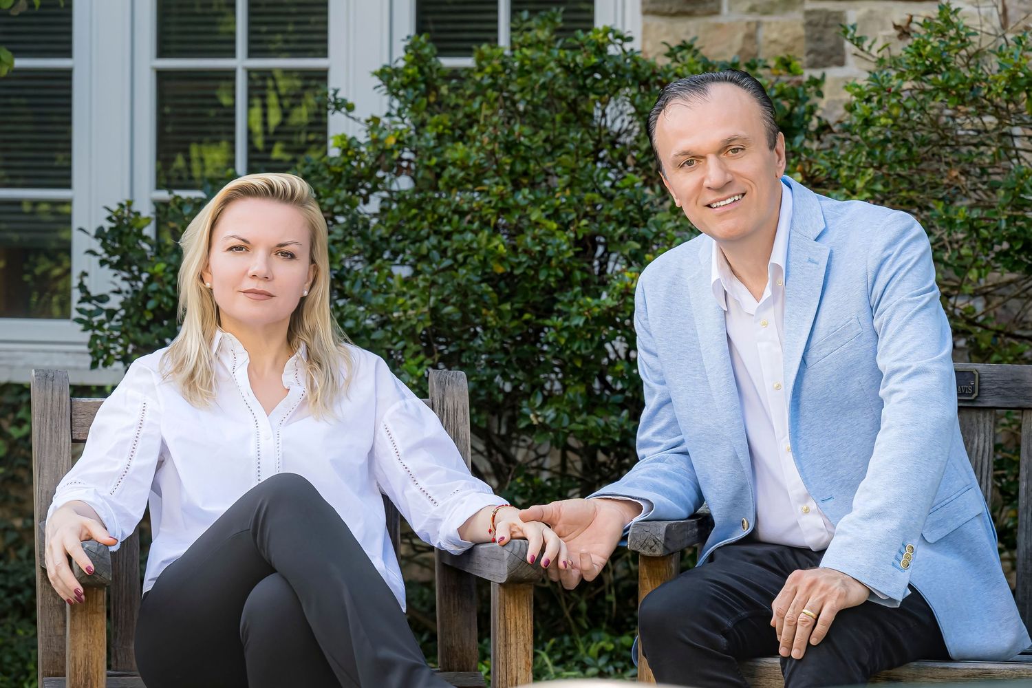 A smiling blonde woman in a white blouse and a man in a light blue blazer sit together holding hands on a wooden bench outdoors.