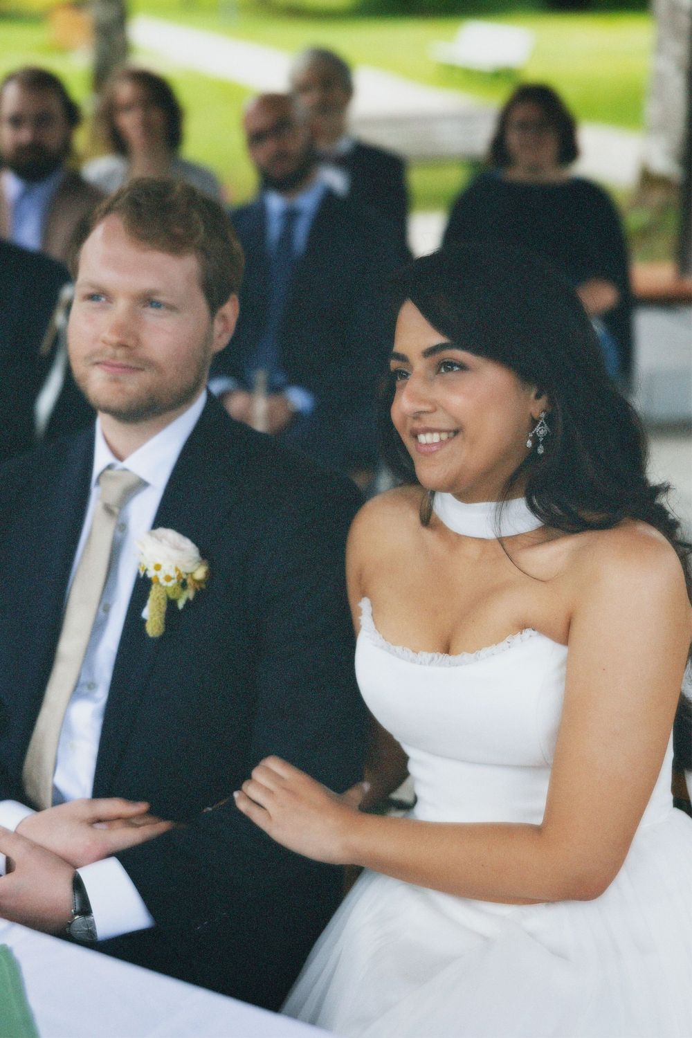 Smiling bride in a white strapless gown sits beside a groom in a dark suit with a boutonniere at an outdoor wedding ceremony with guests.