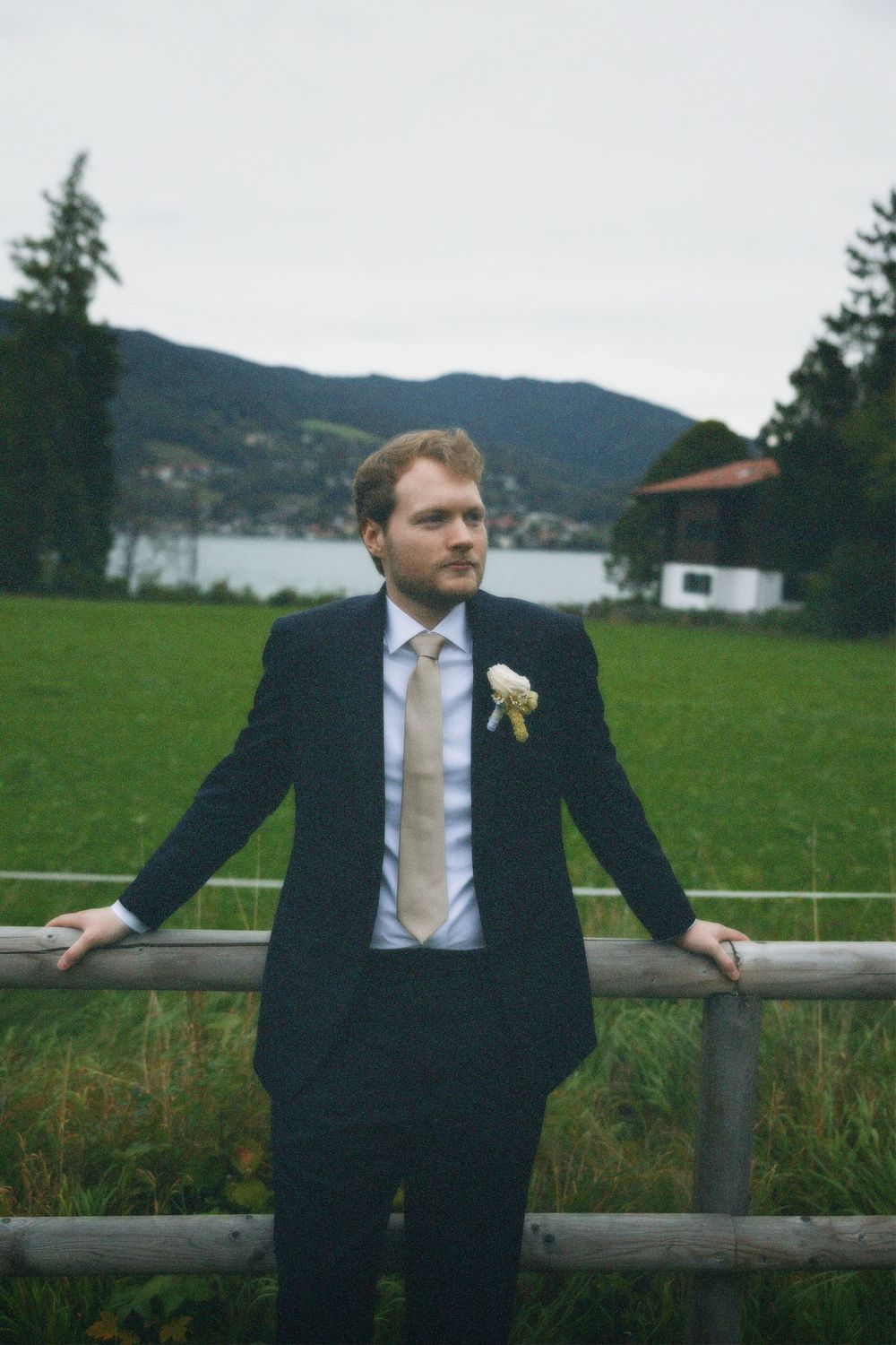 Groom in a navy suit and beige tie with a white boutonniere leans on a wooden fence before a green meadow, lake and mountains.
