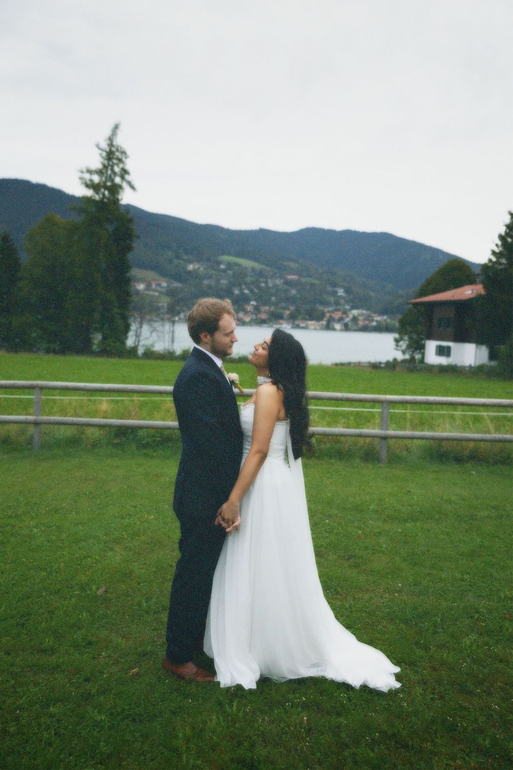 Bride in a flowing white gown and groom in a navy suit embrace on a green lawn with a lake and mountains behind.