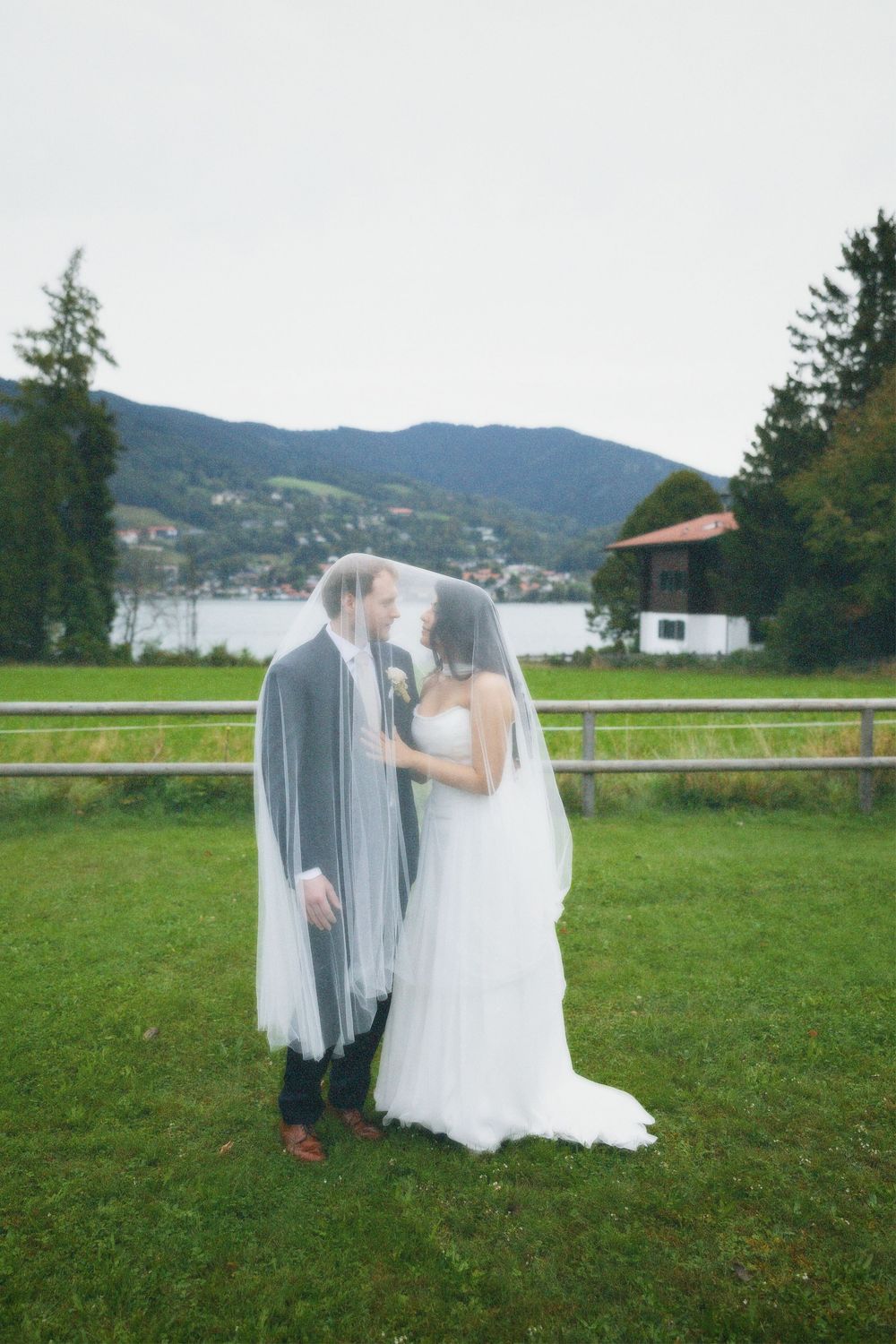 Bride and groom share a quiet moment under a sheer veil on a green lawn with a lake and mountains in the background.