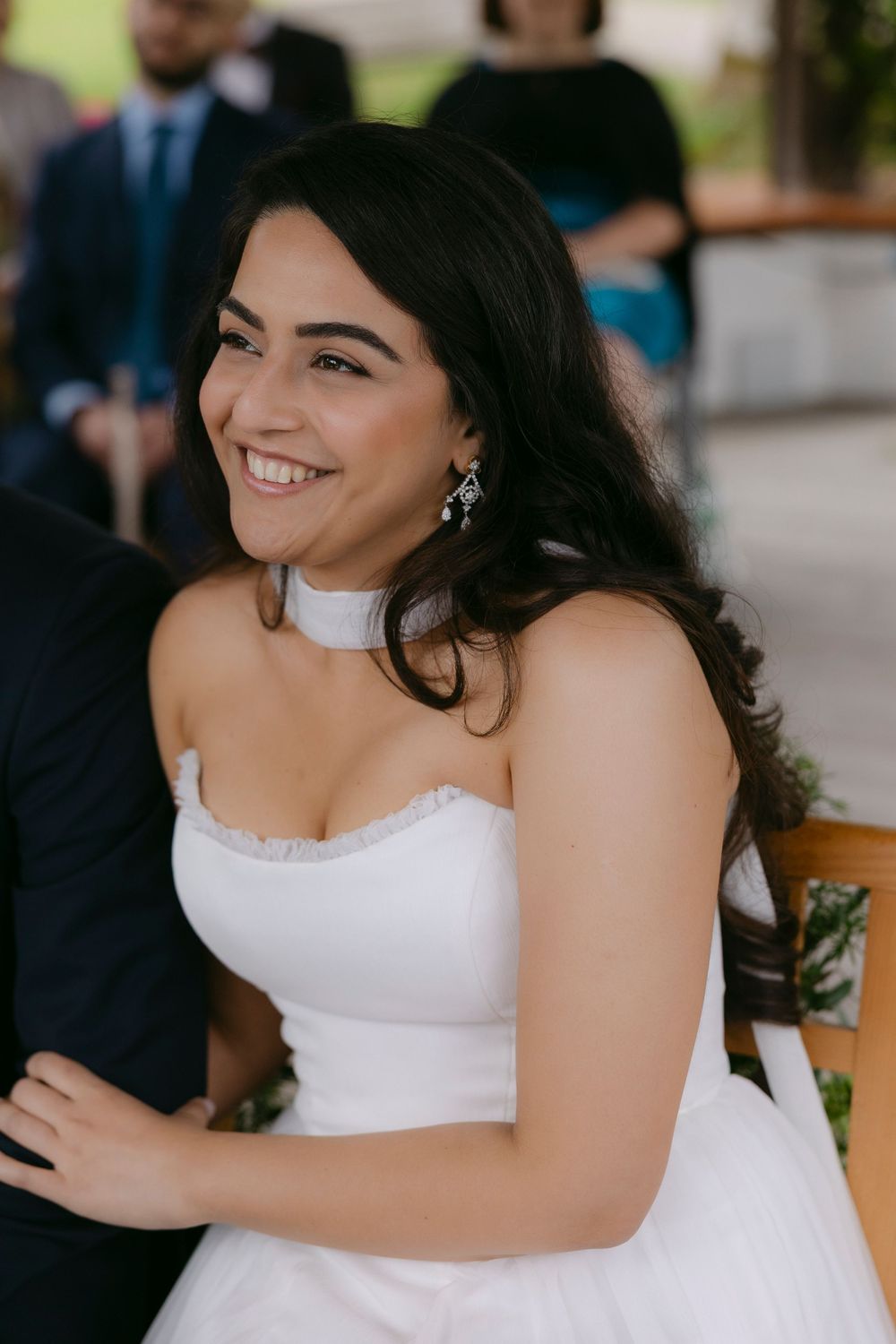 Smiling bride in a strapless white gown and drop earrings laughs during an outdoor wedding ceremony with guests behind her.