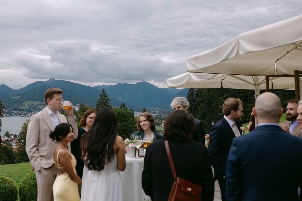 Wedding guests in formal attire chat under white umbrellas on a terrace overlooking a lake and cloudy mountain landscape.