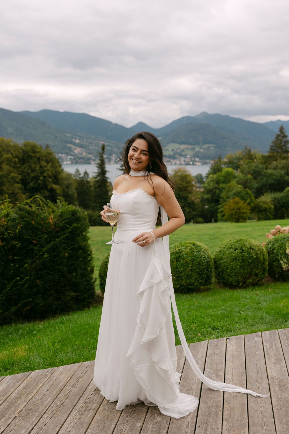 Smiling bride in a flowing white strapless gown holds a champagne glass on a wooden deck overlooking a lake and mountains.