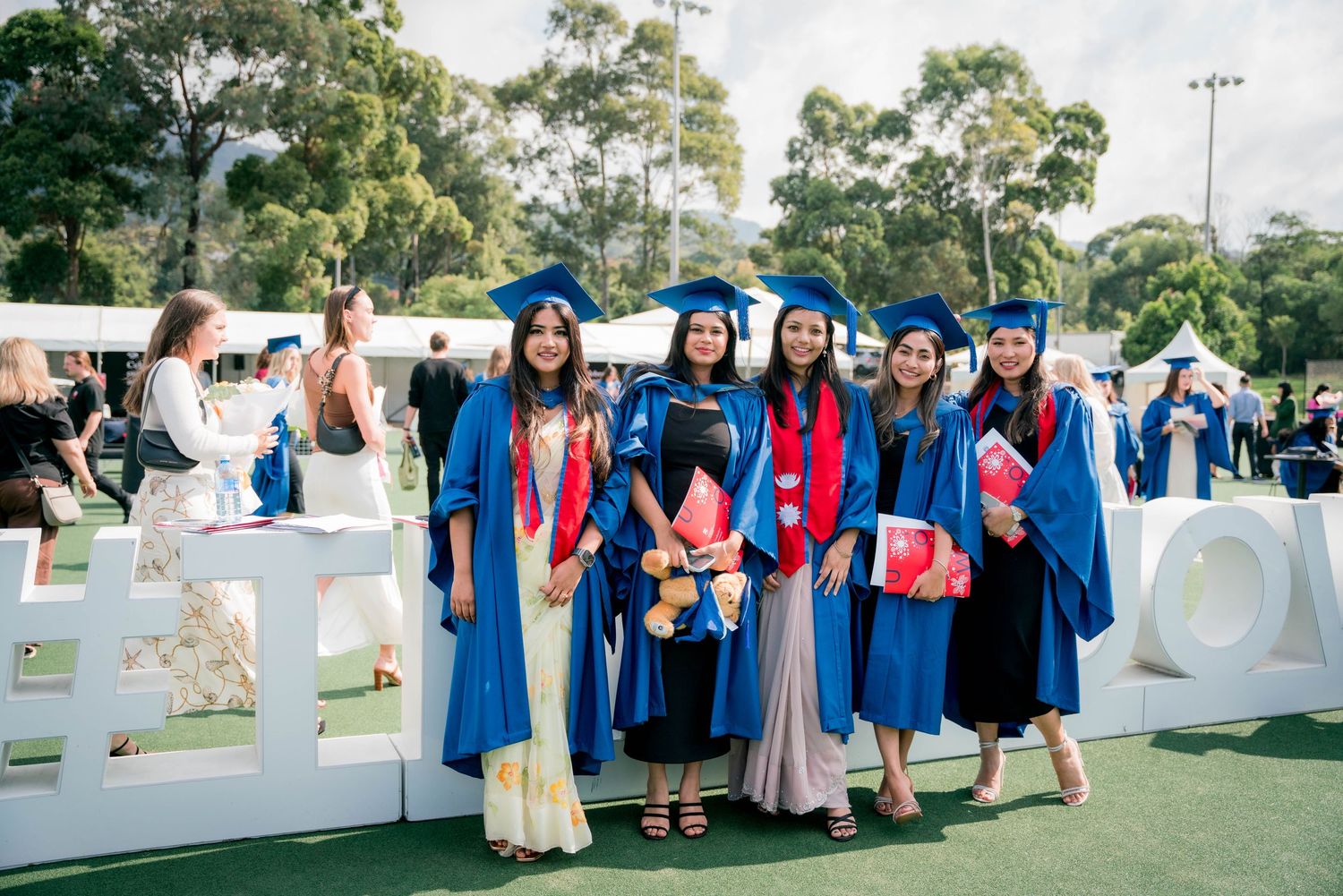 Five female graduates in blue caps and gowns with red stoles pose together on a grassy lawn at an outdoor graduation ceremony.