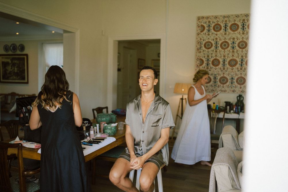 Bridal party getting ready in a bright room as a smiling man in a satin robe sits on a chair while two women stand nearby.