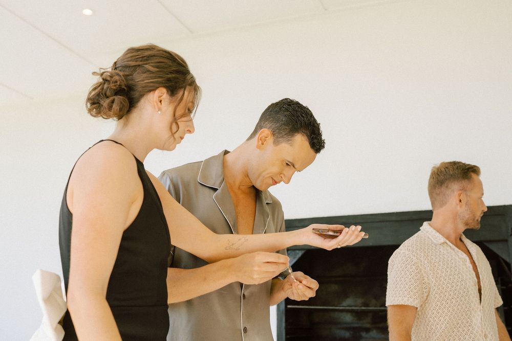 A woman in a black dress and a man in a gray shirt examine a small object together while another man looks on in a bright room.