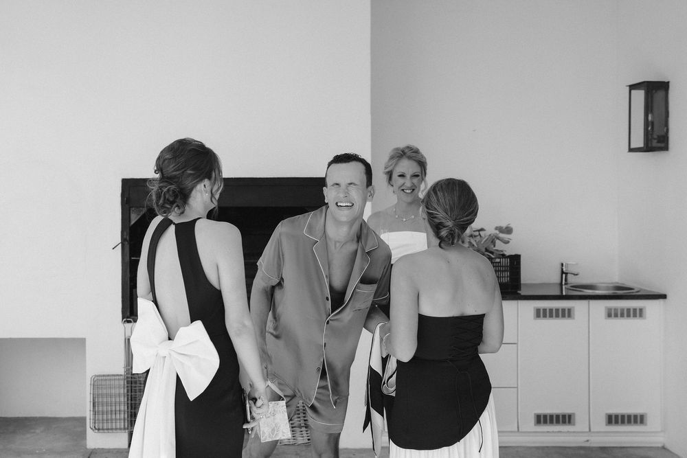 Black and white photo of a laughing man in an open shirt greeting two women in elegant dresses with bow details in a bright room.