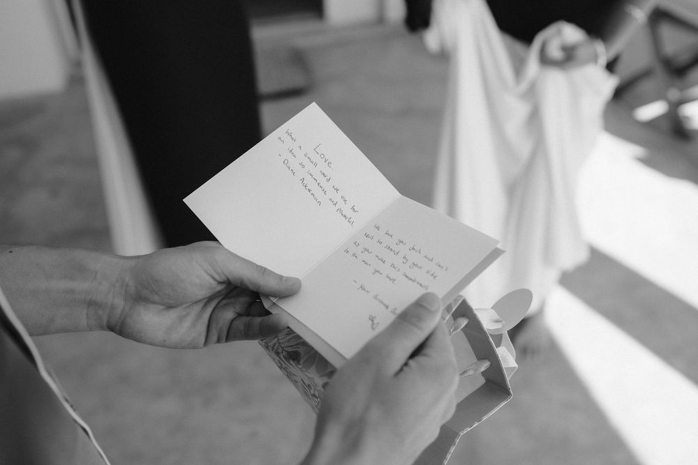 Black and white photo of a person holding an open handwritten letter in both hands, with soft sunlight and blurred fabric in the background.